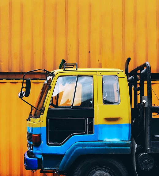 Zijaanzicht van de cabine van een geel-blauwe vrachtwagen, geparkeerd voor een grote, geribbelde oranje scheepscontainer
