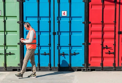 Een logistiek medewerker in een oranje veiligheidsvest loopt met een klembord langs een rij felgekleurde groene, blauwe en rode zeecontainers