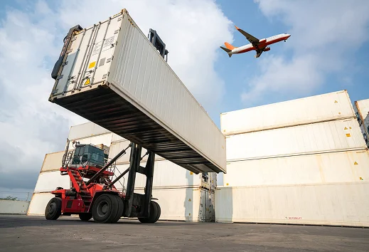 Een rode containerheftruck tilt een grote witte zeecontainer op in een logistieke terminal, terwijl er een passagiersvliegtuig in de blauwe lucht erboven vliegt