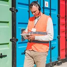 Een close-up van een logistiek medewerker met een headset en een oranje veiligheidsvest, die aantekeningen maakt op een klembord voor kleurrijke zeecontainers