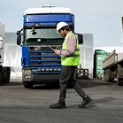 Een logistiek medewerker met een witte helm en een reflecterend vest loopt met een klembord langs een grote blauwe vrachtwagen op een transportterrein